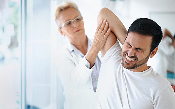 Closeup front angle view of a late 50's female doctor examining a male athlete with some shoulder pain. She's rotating and twisting his shoulder joint and trying to determine which tendons have been damaged. The patient has a painful grimace on his face.