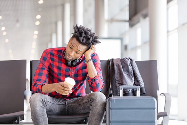 Bored young african american man sitting on chair with suitcase in airport lounge and waiting for flight.